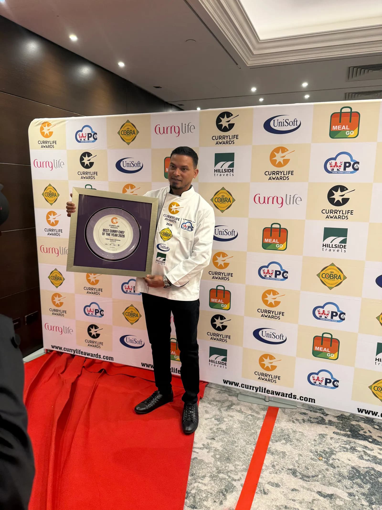 A man in a chef's jacket holds a 'Best Curry Chef of the Year 2020' award plaque in front of a branded backdrop.