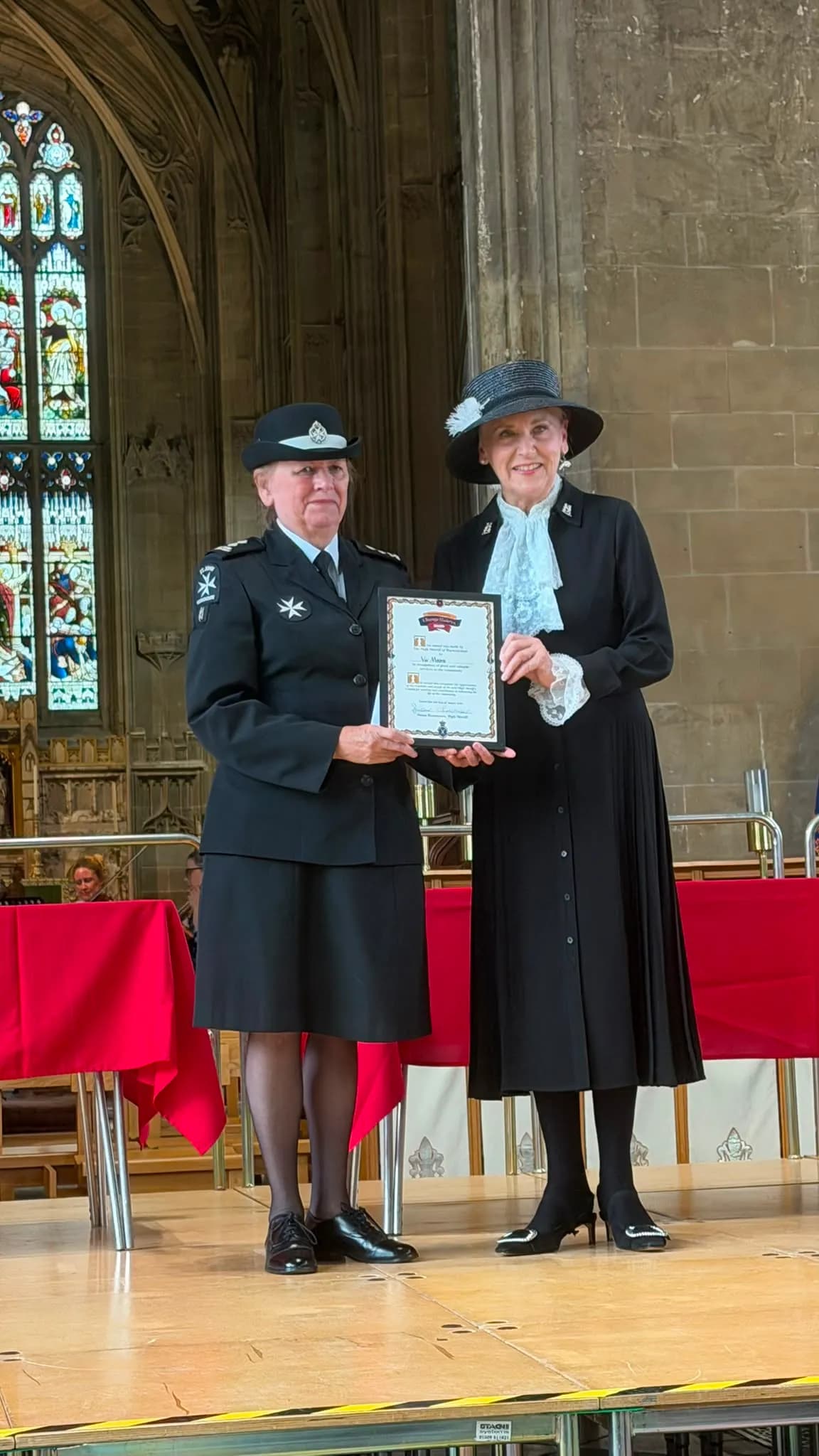 Two women stand on a stage with a stained glass window in the background, one in a uniform holding a framed certificate with the other woman in a black dress and hat.